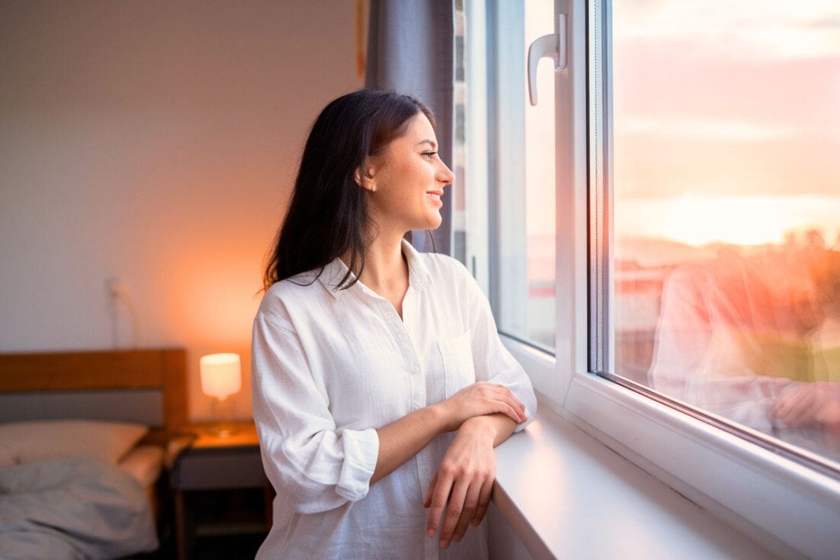 A Happy Woman Looking Out Her Bedroom Window At A Peaceful Sunrise Min